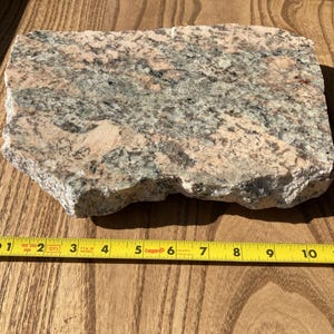 May include: A rectangular piece of granite with a rough, natural edge. The stone features a mix of pink, gray, and black colors. A yellow measuring tape is placed next to the stone, showing a length of approximately 11 inches.