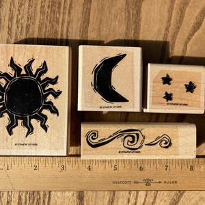 May include: Four wooden rubber stamps featuring black ink designs of a sun, a crescent moon, three stars, and swirling waves. Each stamp is marked with the text Stampin' Up! 1995. A wooden ruler is positioned below the stamps for scale.