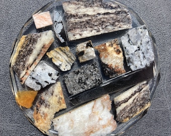 May include: A clear, faceted coaster with an assortment of rock samples. The rocks are in shades of white, grey, black, and orange, showing different textures and patterns. The coaster is circular and rests on a dark surface.