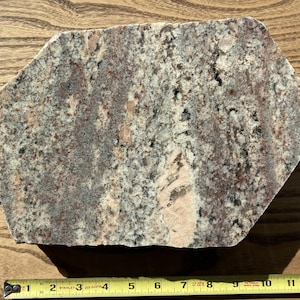 Puede incluir: Una losa de granito de forma aproximadamente hexagonal con una mezcla de tonos grises, rosas y burdeos. La piedra tiene una superficie texturizada y está colocada sobre un fondo de madera. Una cinta métrica amarilla está en la parte inferior.
