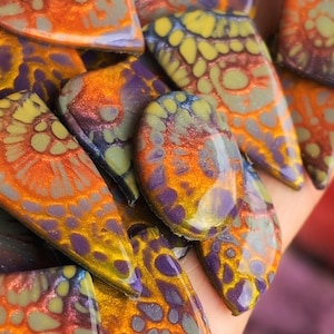 Può includere: Primo piano di una collezione di cabochon colorati con motivi astratti. I cabochon presentano un mix di tonalità arancioni, viola, gialle e verdi con una finitura lucida. Le forme variano, suggerendo che sono destinati alla creazione di gioielli.