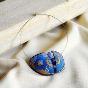 Può includere: Una collana con pendente blu e oro dal design unico. Il pendente presenta una forma geometrica con motivi intricati e una piccola pietra blu rotonda. La collana ha un filo sottile color oro.