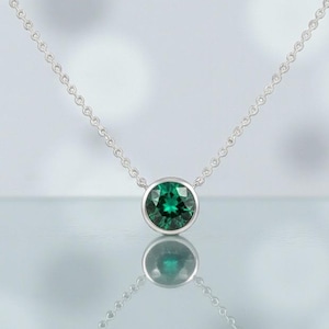 May include: A silver necklace featuring a round, emerald-green gemstone pendant. The gemstone is set in a bezel setting and hangs from a delicate chain. The necklace is displayed on a reflective surface.