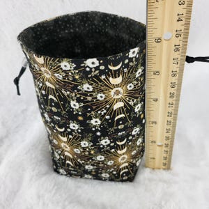 May include: A black fabric drawstring bag with a gold and white moon and star pattern. The bag is approximately 5 inches tall.