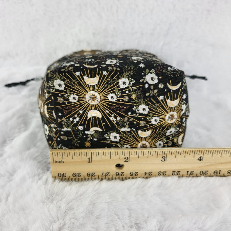 May include: Black drawstring bag with a gold and white floral pattern featuring moons and stars. The bag is approximately 4 inches wide.
