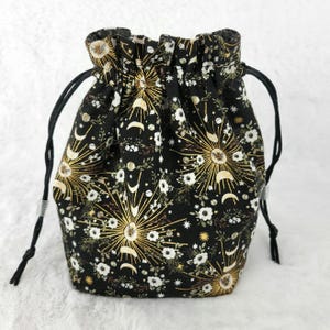 May include: A black drawstring bag with a gold and white floral pattern featuring moons and suns. The bag has a black drawstring closure and is made of fabric.