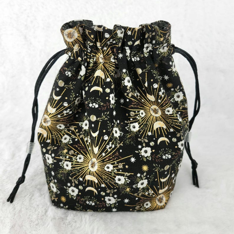 May include: A black drawstring bag with a gold and white floral pattern featuring moons and suns. The bag has a black drawstring closure and is made of fabric.