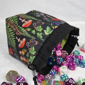 May include: A black drawstring bag with a green and black floral print featuring mushrooms, butterflies, and ferns. The bag is filled with colorful dice.