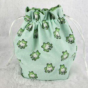 May include: A light green drawstring bag with a white drawstring. The bag is covered in a repeating pattern of green frogs.