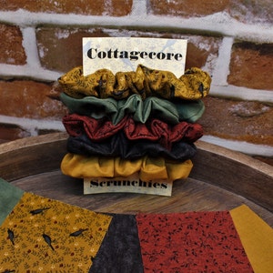 May include: A stack of five fabric scrunchies in various autumnal colors and patterns, including black, green, red, and yellow. The scrunchies are arranged on a wooden surface with a sign that reads "Cottagecore Scrunchies".