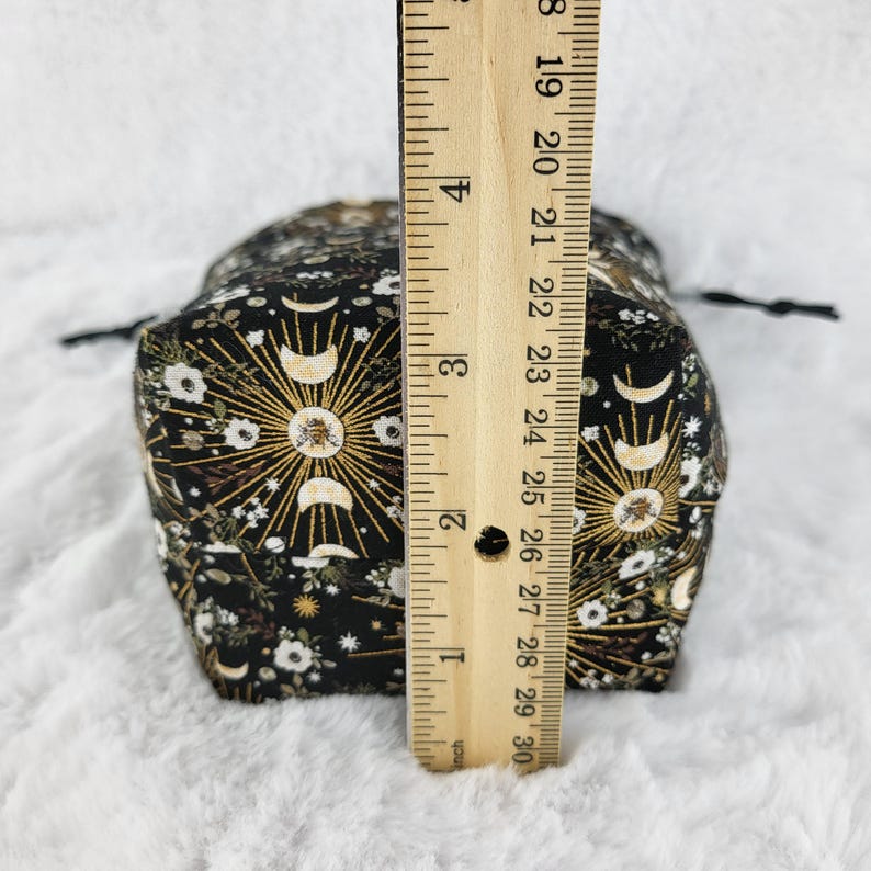 May include: A black fabric drawstring bag with a gold and white moon and sun print. The bag is approximately 4 inches tall.