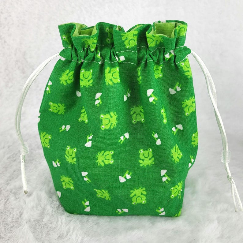 May include: Green drawstring bag with a white frog and mushroom pattern. The bag has white drawstrings.