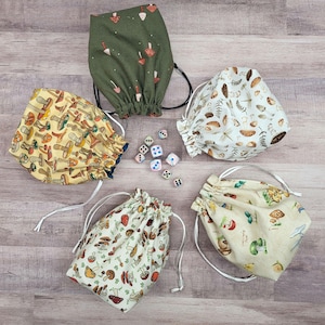May include: Four fabric drawstring pouches featuring various mushroom patterns in olive green, yellow, cream, and off-white colors, arranged around a small collection of six-sided dice with different colors and numerical pips on their faces.