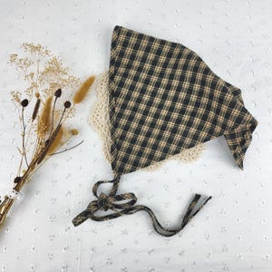 May include: A black and tan plaid bonnet with a cream-colored lace trim and matching ties. The bonnet is made of a soft fabric and is designed to be worn on the head. The ties are used to secure the bonnet.