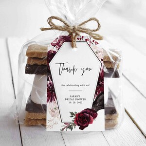May include: A white thank you tag with black text that reads "Thank you for celebrating with us!"  Sarah's Bridal Shower 10.29.2022. The tag is decorated with a floral design featuring burgundy and pink flowers and greenery. The tag is tied to a clear plastic bag containing a s'mores kit.