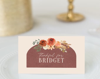 Puede incluir: Una tarjeta de sitio rectangular con fondo crema y una forma de arco de color granate oscuro. El texto "Thankful for BRIDGET" está impreso en blanco, coronado con una ilustración en acuarela de flores naranjas, rojas y marrones con hojas verdes.