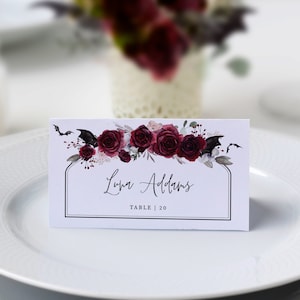 Op de afbeelding: Een witte naambordje met een zwarte rand, met de naam "Luna Addams" en tafelnummer 20. De kaart is versierd met bordeauxrode rozen, donkere bladeren en vleermuis silhouetten. De kaart ligt op een wit bord.