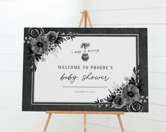 May include: A black and white baby shower sign on a wooden easel. The sign reads "Welcome to Phoebe's baby shower" with floral accents. The date "October 29, 2023" is also displayed. The sign is framed with a dark gray border.
