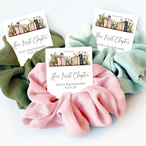 May include: Three satin scrunchies in olive green, light pink, and seafoam green, each with a small card. The cards read "Her Next Chapter" and "Julie's Bachelorette 4.25.26" with a floral and book design. These are ideal for a bachelorette party.
