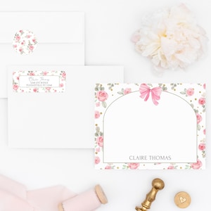 May include: A stationery set featuring a floral design with pink roses and green leaves. The set includes envelopes, labels, and a card with a pink bow. A gold wax seal and spool of pink ribbon are also present.