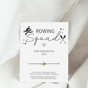May include: A teal blue friendship bracelet with a gold star charm. The card reads "ROWING Squad" with a heart and two crossed oars. The card also says "TEAM DRAGONETTES 2023" and "CLOSE YOUR EYES AND MAKE A WISH, THEN TIE THIS BRACELET AROUND YOUR WRIST. WHEN THE STRING HAS WORN THROUGH, THE WISH YOU MADE MAY COME TRUE!"