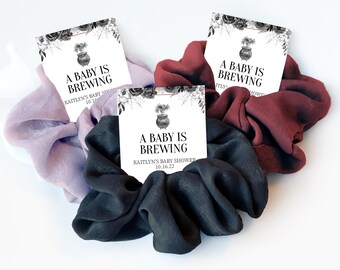May include: Three scrunchies in shades of purple, burgundy, and black, each holding a card. The cards read "A Baby Is Brewing" with a cauldron graphic and "Kaitlyn's Baby Shower 10.16.22". The scrunchies are made of a silky fabric.