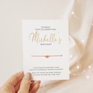 Op de afbeelding: Een roze koordarmband met een gouden hartbedel. De kaart luidt: "Bedankt voor het vieren van Michelle's verjaardag. Sluit je ogen en doe een wens, bind dan deze armband om je pols. Wanneer het koord versleten is, kan je wens uitkomen!"