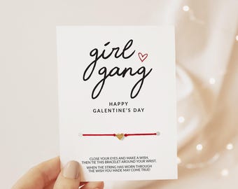 May include: A red string bracelet with a small gold heart charm attached to a white display card. The card features the black text "girl gang" with a red heart, "HAPPY GALENTINE'S DAY", and instructions about making a wish.