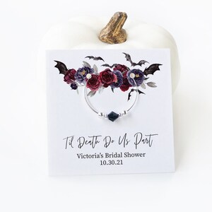 May include: A wine glass charm with a floral and bat design. The charm features a silver ring with a dark green gem and silver beads. The card reads "Til Death Do Us Part" and "Victoria's Bridal Shower 10.30.21".