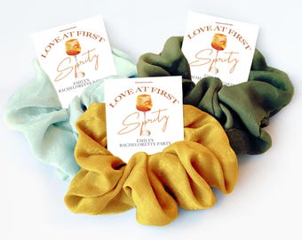 May include: Three fabric hair scrunchies in light blue, mustard yellow, and olive green. Each scrunchie is attached to a white square card featuring an illustration of a spritz cocktail and the text Love at First Spritz Emily's Bachelorette Party.