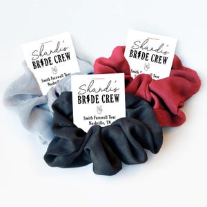 May include: Three fabric hair scrunchies in light blue, dark red, and black. Each is attached to a white card printed with the text Shandi's BRIDE CREW, a hand gesture icon, and the text Smith Farewell Tour Nashville, TN.