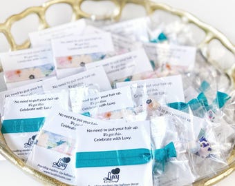May include: A collection of teal elastic hair ties packaged in individual clear plastic sleeves with white cards. Each card features the text: No need to put your hair up. We got this. Celebrate with Luxy. Luxy Balloons logo is printed at the bottom.