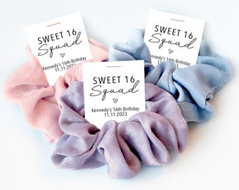 May include: Three fabric hair scrunchies in pastel pink, light blue, and lavender, each attached to a white card printed with the text SWEET 16 Squad, a small heart icon, and the text Kennedy's 16th Birthday 11.11.2023.