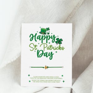 Peut inclure: Une carte blanche avec le texte vert Happy St. Patricks Day, un chapeau de lutin vert et des trèfles. Un bracelet en fil vert avec une petite étoile dorée est fixé sur la carte. En bas, le texte indique : CLOSE YOUR EYES AND MAKE A WISH, THEN TIE THIS BRACELET AROUND YOUR WRIST. WHEN THE STRING HAS WORN THROUGH, THE WISH YOU MADE MAY COME TRUE!