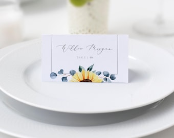 May include: A rectangular white place card featuring a thin black border, the printed name Willow Morgan, the text TABLE | 10, and a watercolour illustration of a yellow sunflower with blue-green eucalyptus leaves at the bottom.