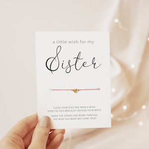Op de afbeelding: Een roze armband van touw met een gouden hartbedel. De kaart staat "a little wish for my Sister" en "CLOSE YOUR EYES AND MAKE A WISH, THEN TIE THIS BRACELET AROUND YOUR WRIST. WHEN THE STRING HAS WORN THROUGH, THE WISH YOU MADE MAY COME TRUE!"