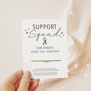 May include: Green string bracelet with a gold heart charm and a card that reads "Support Squad Team Jennifer accept. love. understand Close your eyes and make a wish, then tie this bracelet around your wrist. When the string has worn through, the wish you made may come true!"
