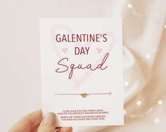 Puede incluir: Una tarjeta blanca con el texto rojo GALENTINE'S DAY Squad, un contorno de corazón rosa claro y una pulsera de hilo rosa fino con un pequeño dije de corazón dorado. La tarjeta incluye instrucciones para pedir un deseo y atar la pulsera.