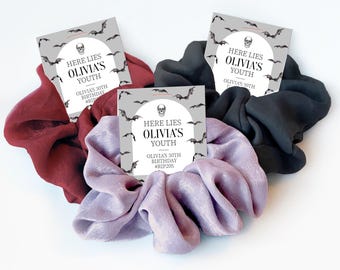 May include: Three fabric hair scrunchies in burgundy, black, and lavender, each attached to a square card featuring a skull icon, flying bats, and the text: HERE LIES OLIVIA'S YOUTH, OLIVIA'S 30TH BIRTHDAY, #RIP20S.