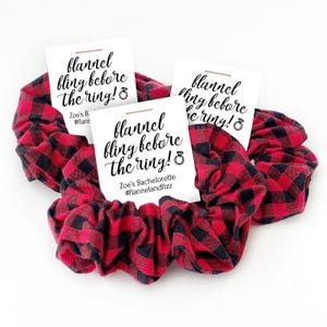 May include: Three red and black buffalo plaid fabric hair scrunchies, each attached to a white card featuring the black text "flannel fling before the ring!" with a ring icon, "Zoe's Bachelorette", and the hashtag "#flannelandfizz".