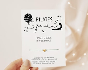 May include: A light blue string bracelet with a small gold heart charm attached to a white display card. The card features black text reading Pilates Squad, Oxygen Studios, Inhale. Exhale, and instructions to make a wish.