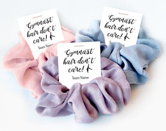 Puede incluir: Tres coleteros de tela en rosa claro, azul claro y lavanda. Cada coletero está sujeto a una tarjeta blanca con el texto negro "Gymnast hair don't care!" y la silueta de un gimnasta, con espacio para escribir el nombre del equipo.