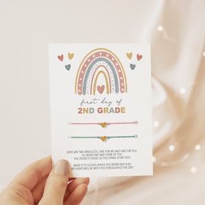 May include: A card with a rainbow illustration and the text "first day of 2ND GRADE". Two bracelets with gold heart charms are attached to the card.
