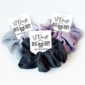 May include: Three fabric hair scrunchies in light blue, lavender, and black. Each scrunchie is attached to a white card featuring a skull and crossbones graphic with the text "Til Death DO US PARTY" and "Amy's Bachelorette" printed in black.