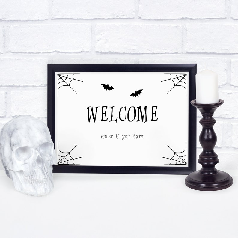 May include: A Halloween-themed framed sign with the word "WELCOME" and the phrase "enter if you dare" in black text. The sign features spiderwebs and bat silhouettes. A white skull and a dark brown candle holder with a white candle are also present.