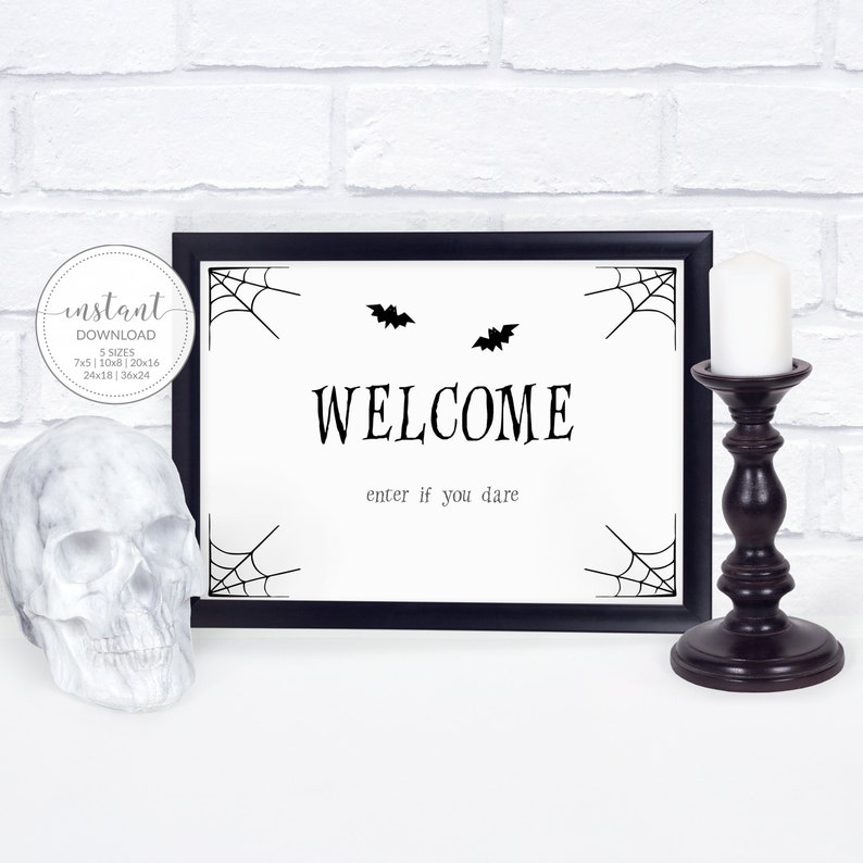 May include: A framed Halloween sign with the word "WELCOME" in a bold font, accompanied by the phrase "enter if you dare." The sign features spiderwebs and bat silhouettes. A white skull and a dark brown candle holder are also visible.