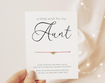 May include: A white card featuring the text 'a little wish for my Aunt' in black script and block lettering. A thin pink string bracelet with a small gold-colored heart charm is attached to the center. The card includes instructions to make a wish.