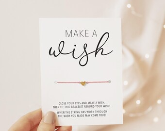 May include: A white rectangular card featuring the black text MAKE A wish and a pink string bracelet with a small gold heart charm. The card includes instructions to close your eyes, make a wish, and tie the bracelet around your wrist.