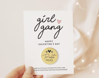 May include: A white rectangular card featuring the black text girl gang with a small red heart, the words HAPPY GALENTINE'S DAY, and a round gold metal token printed with three black hearts and the words Pocket HUG.