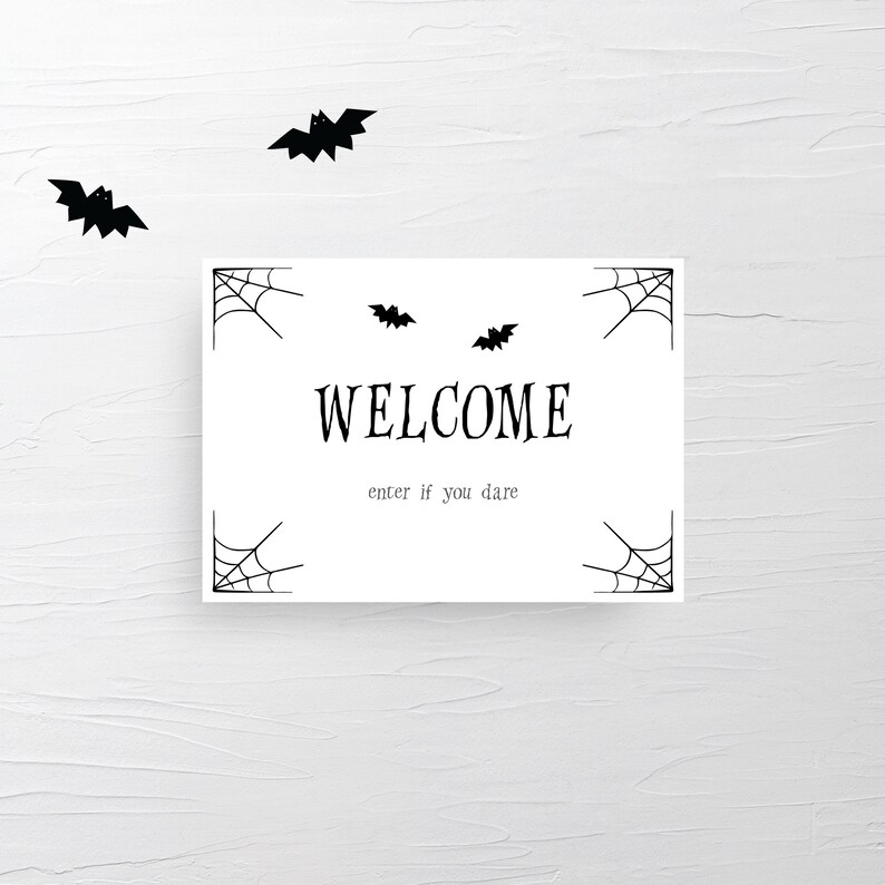 May include: A white rectangular sign with the word "WELCOME" in a bold, black font, and the phrase "enter if you dare" below it. The sign has spiderwebs in each corner and is decorated with black bat silhouettes. Halloween-themed decor.